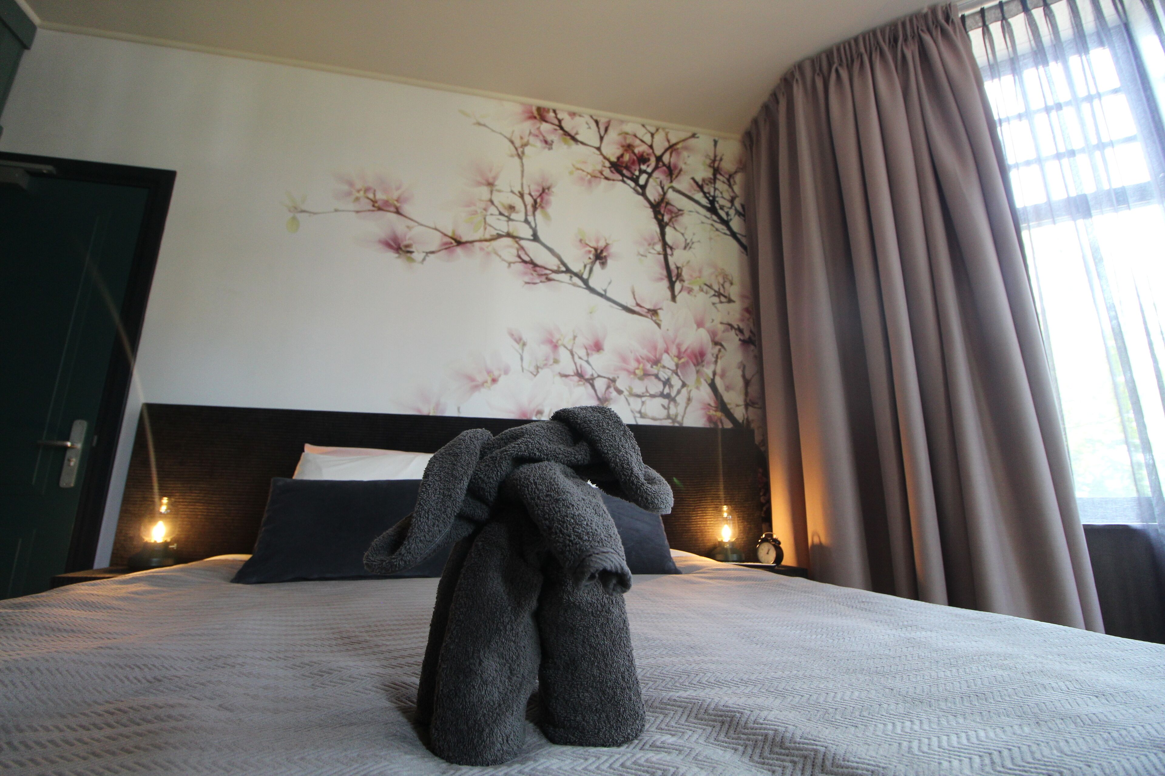 Une chambre d’hôtel avec un lit, un mur orné d’une fresque florale, une fenêtre avec des rideaux et une table de chevet avec une lampe.