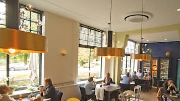 A restaurant interior with multiple dining tables and chairs, large windows with blinds, hanging pendant lights, and a visible outdoor view.