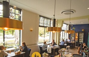 L’intérieur d’un restaurant présente de nombreuses tables et chaises, de grandes fenêtres équipées de stores, des luminaires suspendus et offre une vue dégagée sur l’extérieur.