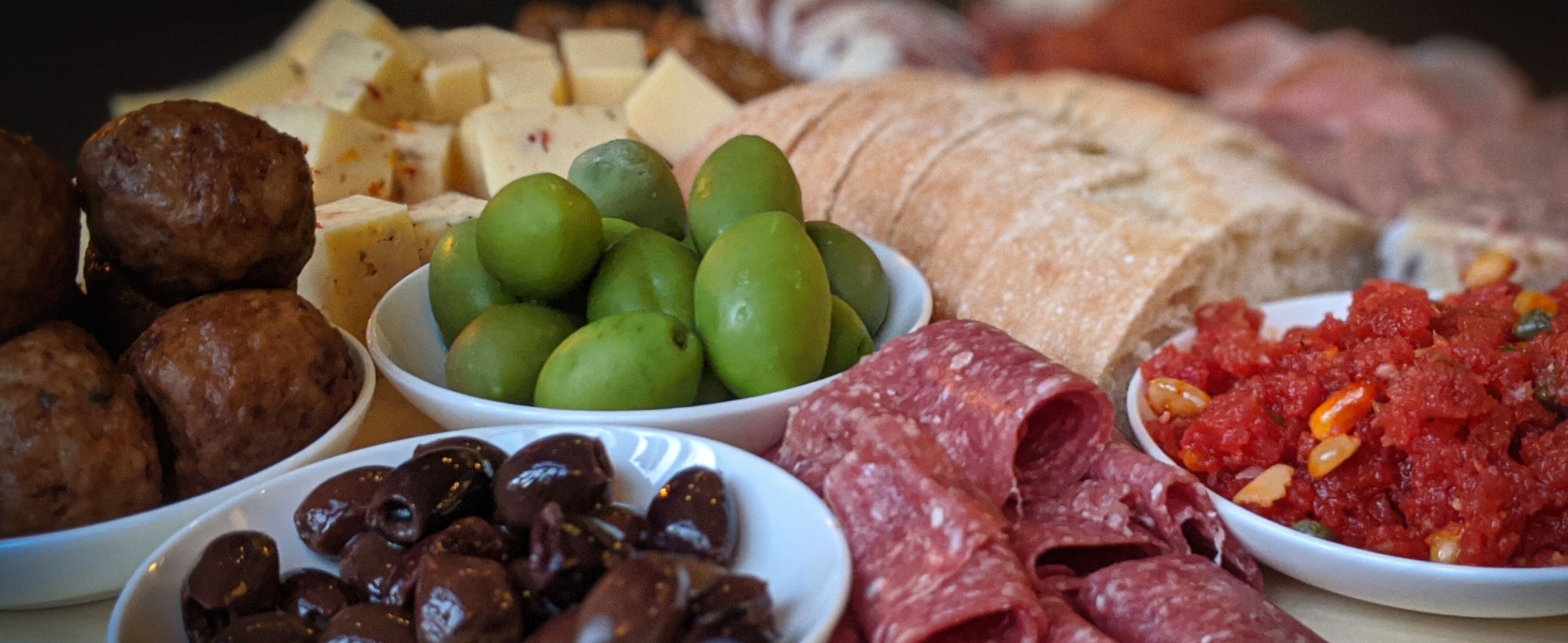 Diversos productos alimenticios como aceitunas, queso, embutidos y una barra de pan.