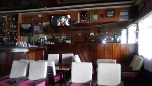 A bar with wooden paneling, a television mounted on the wall, and several white cushioned chairs. - Sundowner Lodge (Nakuru)