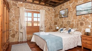 Une chambre avec un mur en pierre, des poutres en bois au plafond, un lit recouvert d’une couverture bleue, une table de chevet avec une lampe et une fenêtre avec des rideaux.