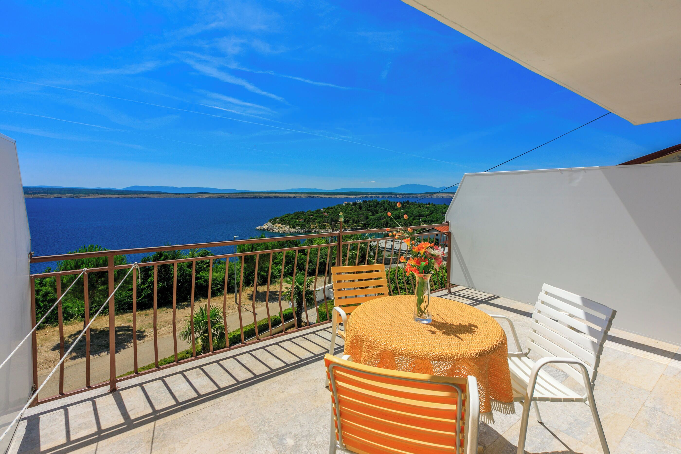 Ein Balkon mit Tisch und Stühlen, mit Blick auf ein Gewässer und ein entferntes Land.
