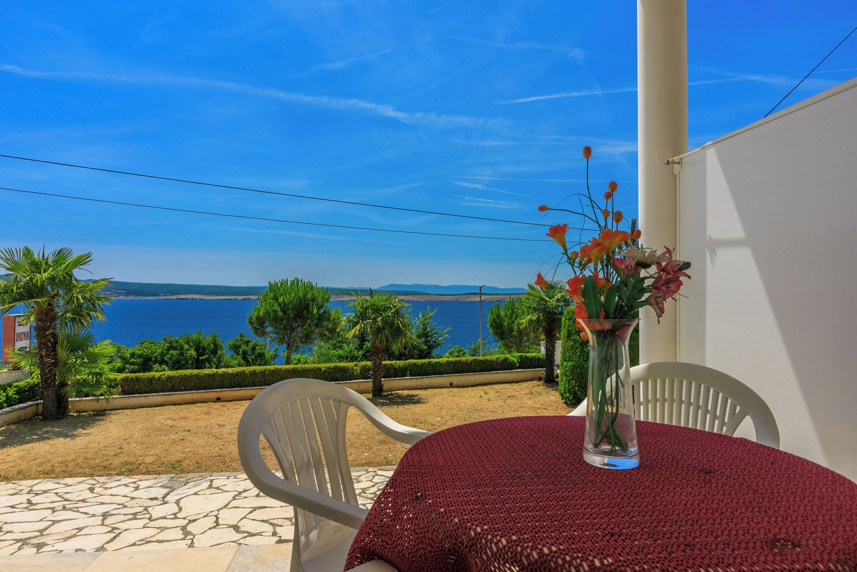 A terrace with a table and chairs, a vase with flowers, and a view of the sea and mountains.