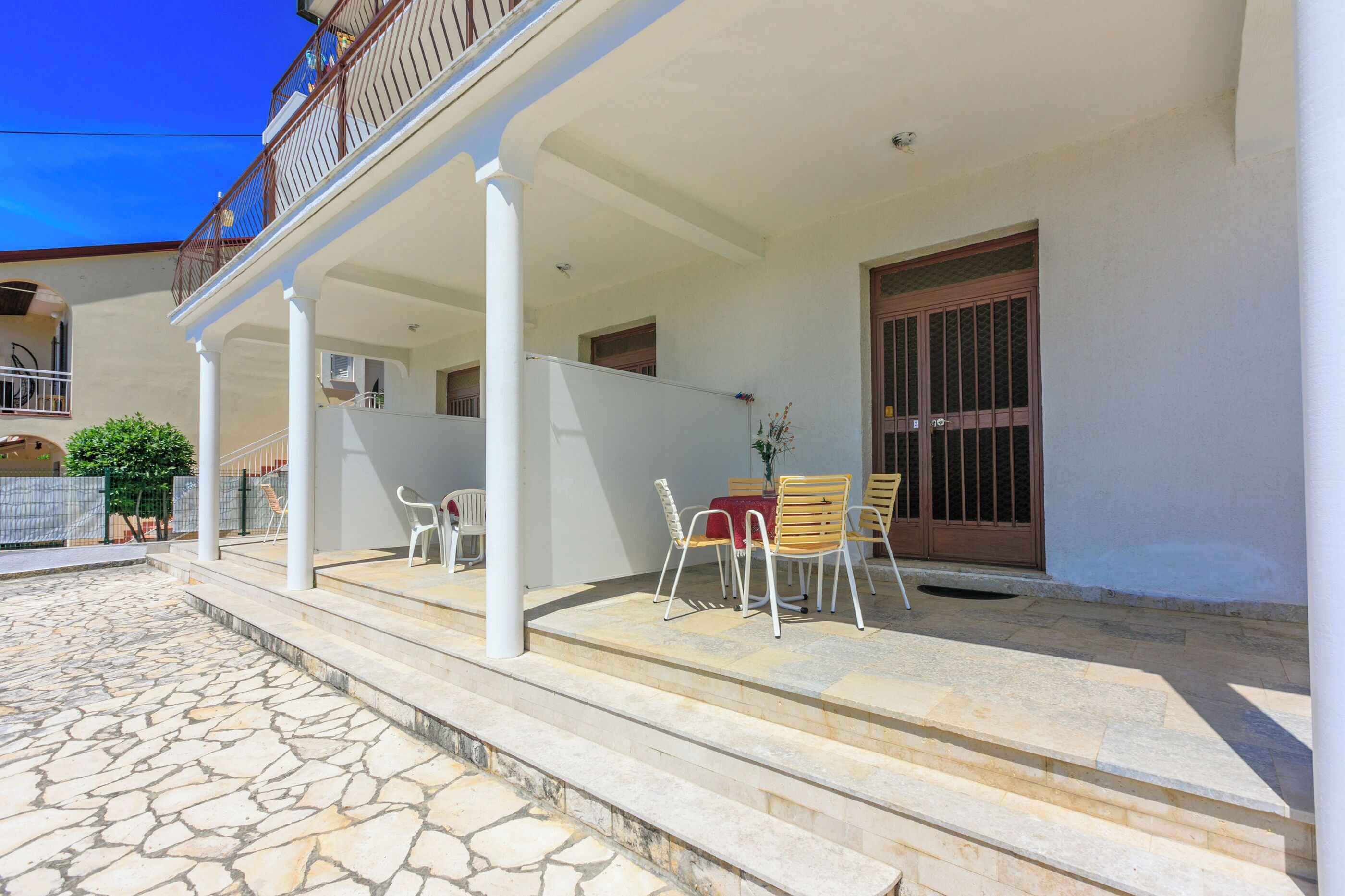 A covered outdoor area with stone flooring, white columns, and a table with chairs.
