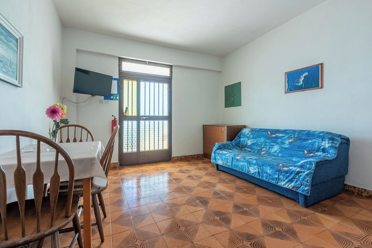 A room with a dining table, chairs, a sofa, a TV mounted on the wall, and a patterned floor.