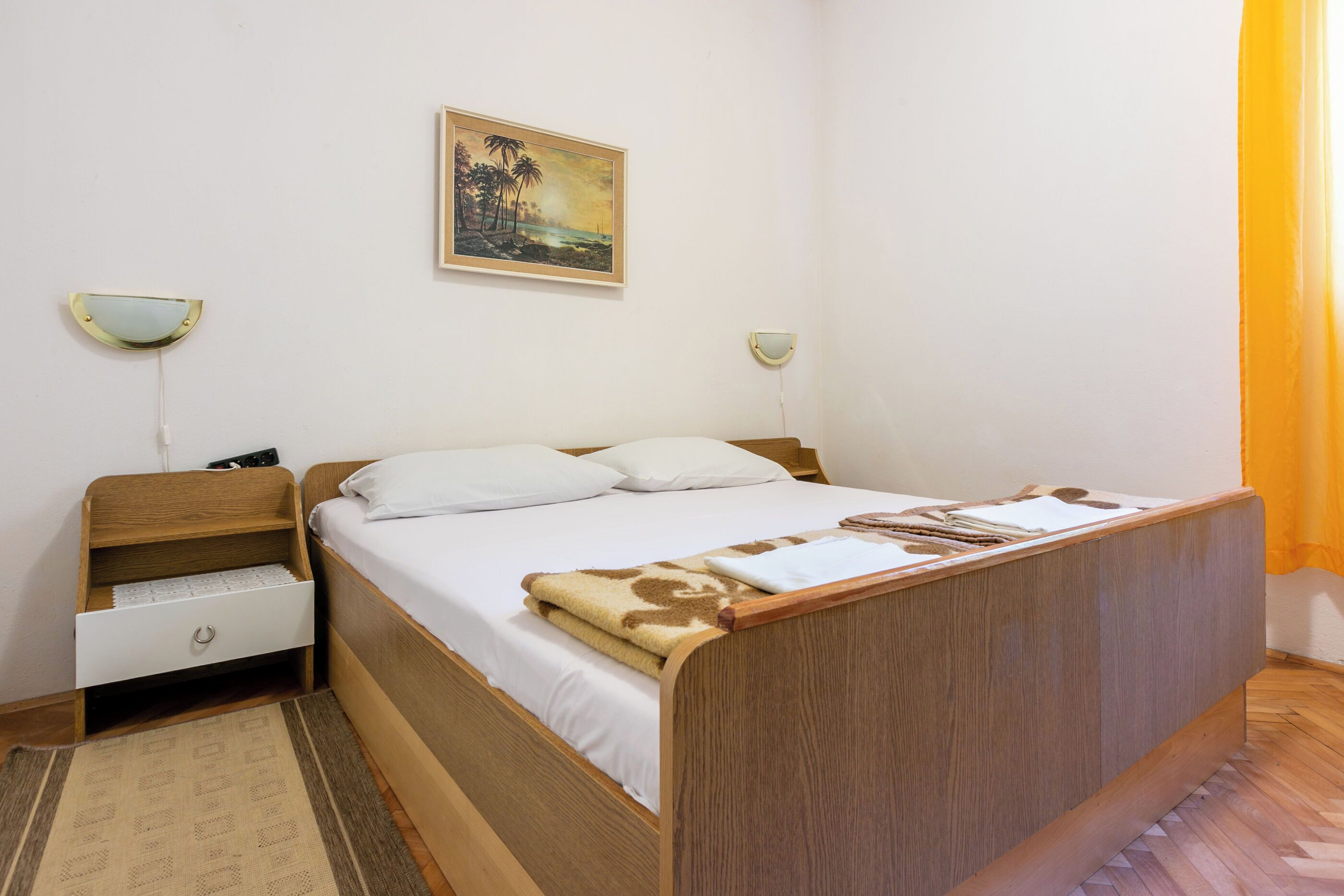 A bedroom with a wooden bed, white bedding, a small bedside table with a lamp, and a painting on the wall.