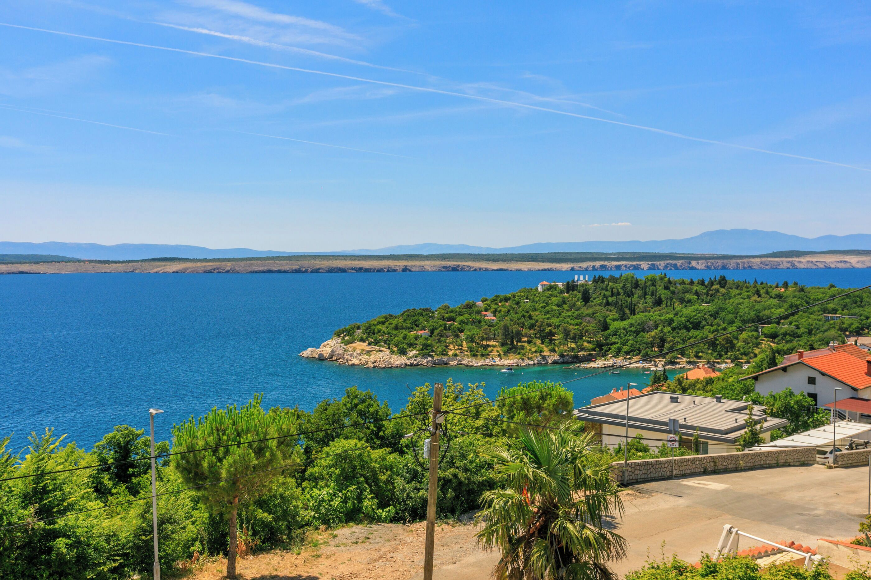 A coastal view with a clear blue sea, a small island with greenery, and a building with a red roof.