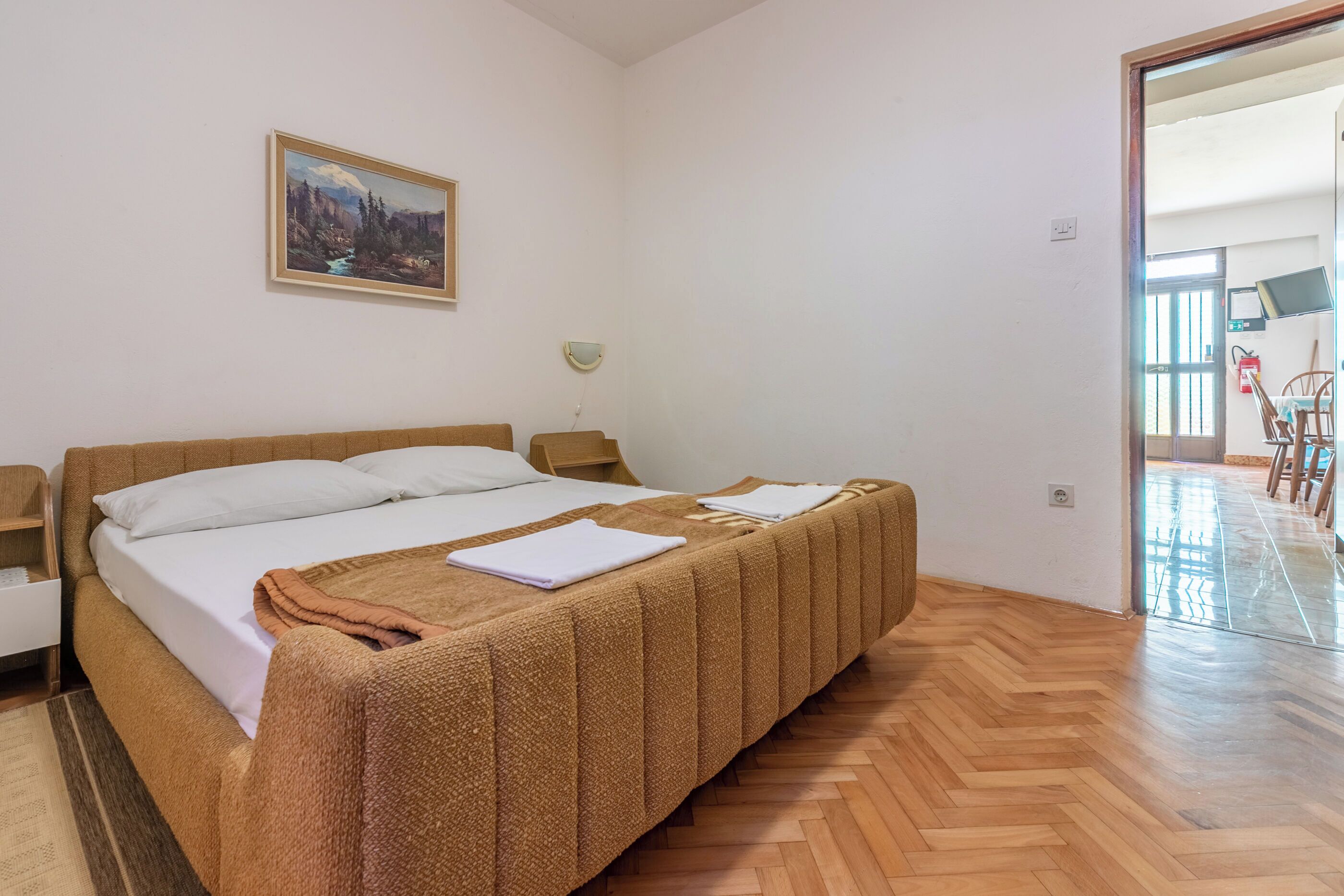 A hotel room with a large bed, a painting on the wall, a bedside table, and a wooden floor.