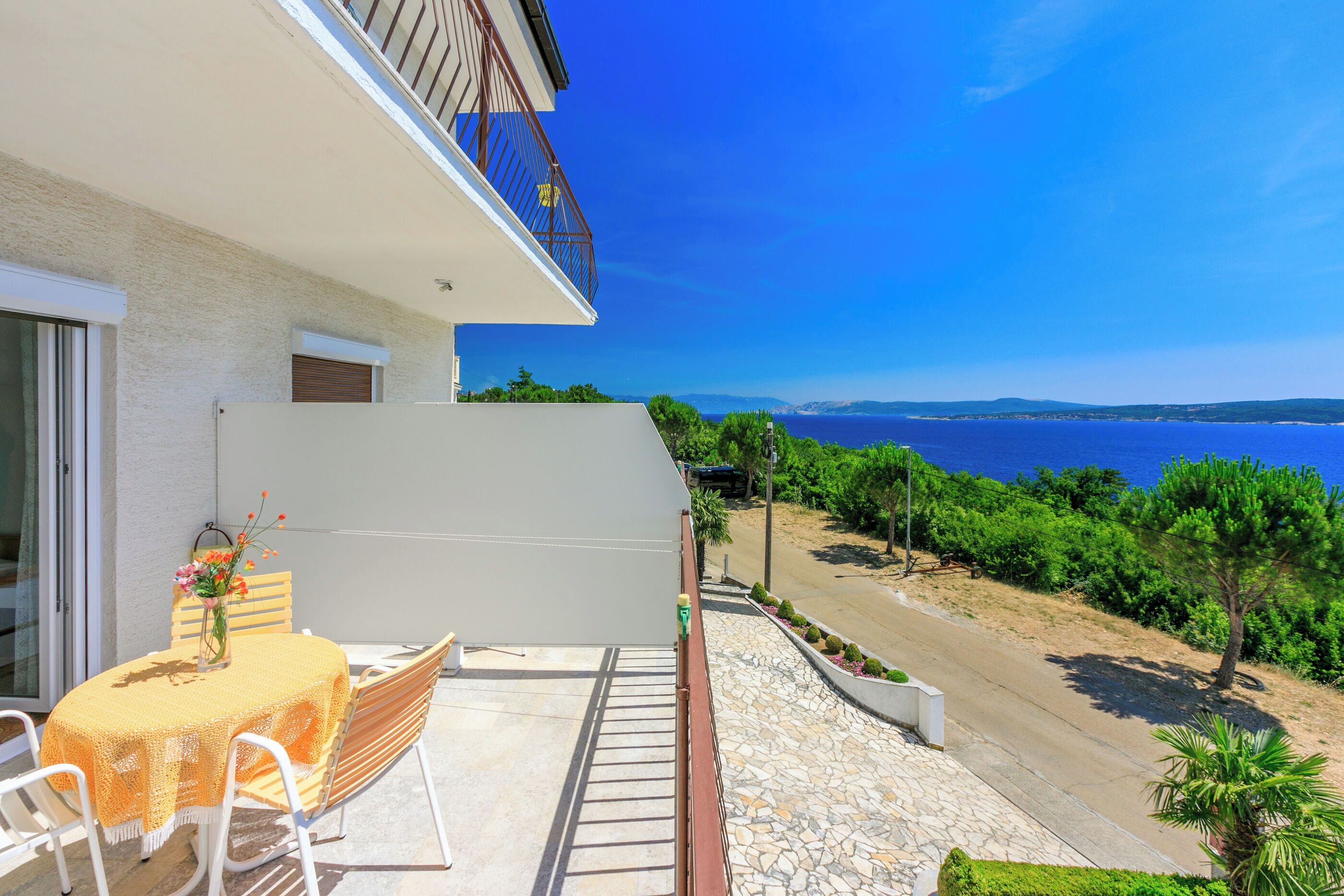 A balcony with a table and chairs, overlooking a coastal road and sea.