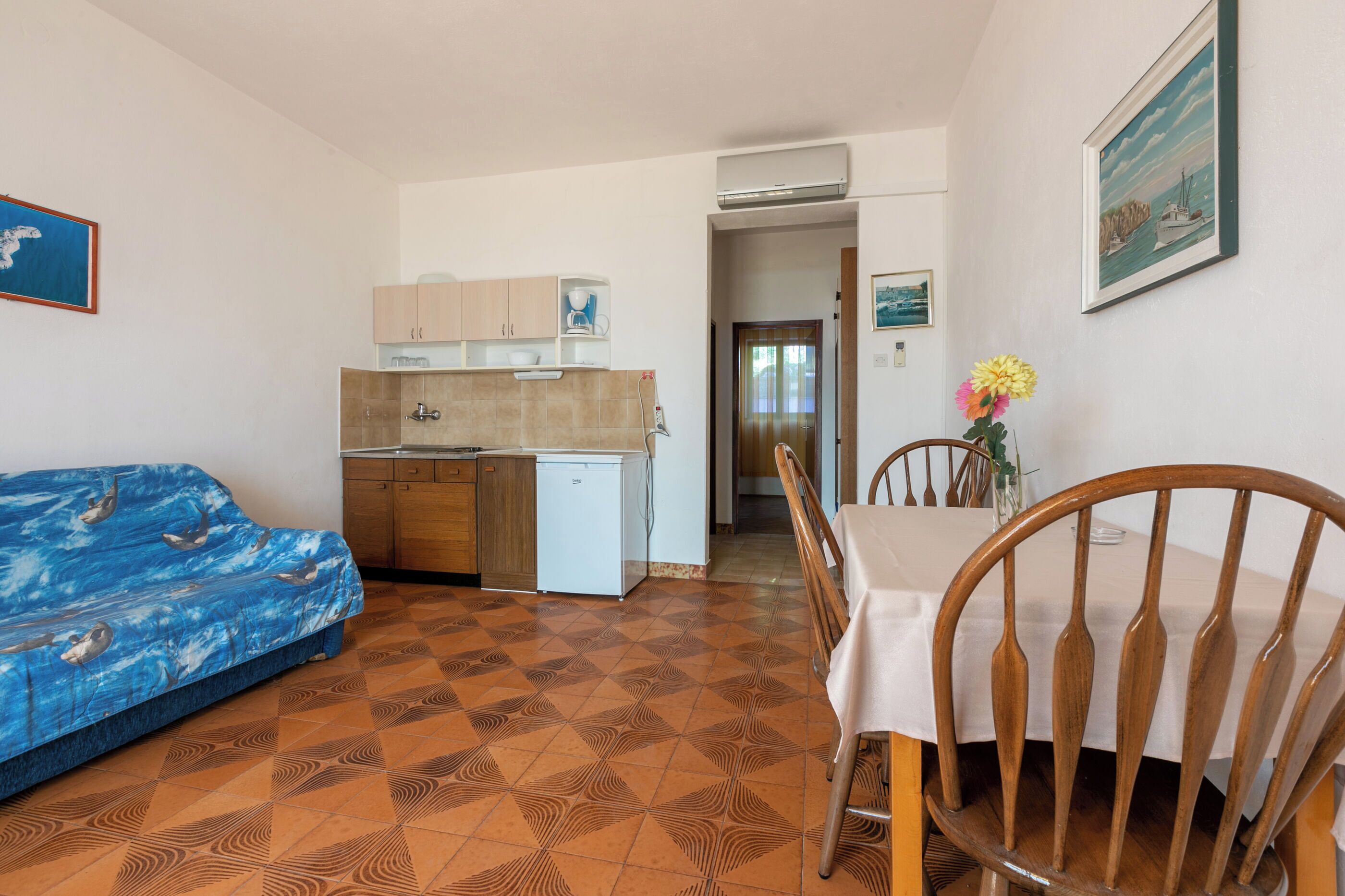 Un espacio habitable compacto con kitchenette, una zona de comedor con mesa y sillas de madera, y un sofá con funda de color azul con motivos marinos.
