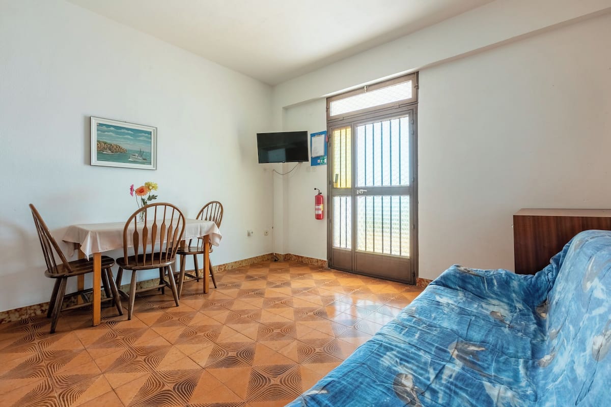 A room with a tiled floor, a blue sofa, a wooden table with chairs, a television mounted on the wall, and a window with bars.