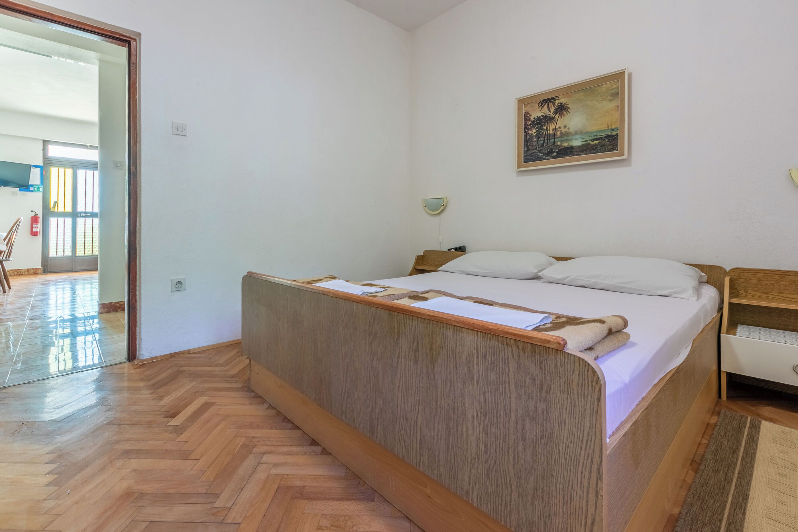 Une chambre avec un lit en bois, une table de chevet blanche, un tableau au mur et un parquet à motifs en chevrons.