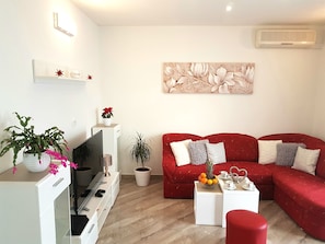 A modern living room with a red sofa, a white coffee table, and a flat-screen TV. There are potted plants and a wall-mounted artwork. - Apartments Filip (Omis)