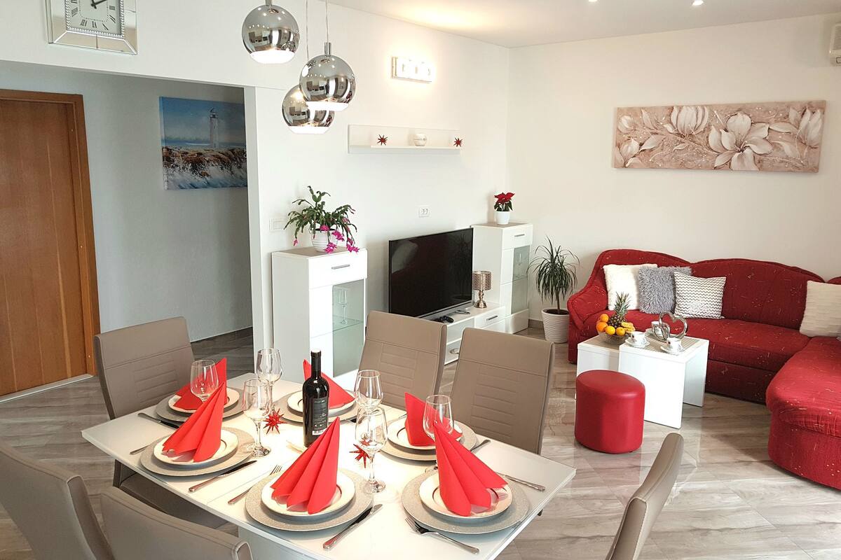 A modern living room with a red sofa, a dining area with a set table, and a TV in the background.