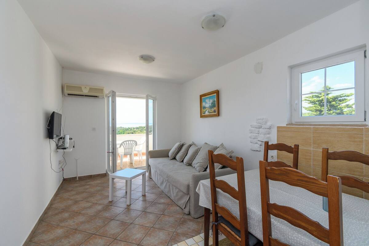 Una habitación con sofá, una silla de madera, una mesita, un televisor en la pared y una ventana con vistas.