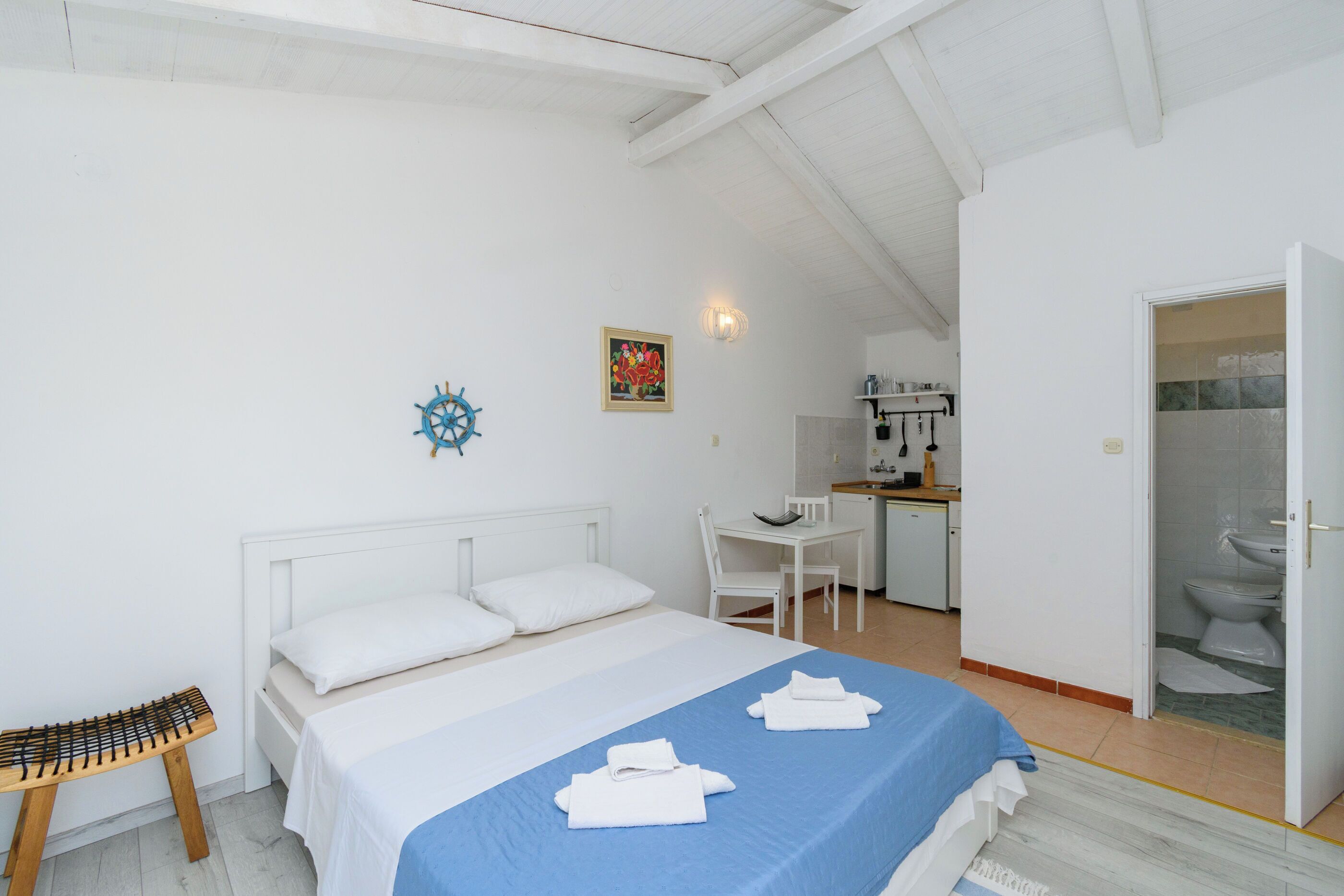 A bedroom with a bed, a desk, and a chair, a small stool, and a bathroom visible through an open door.