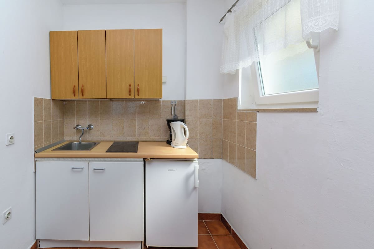 Eine kompakte Küche mit Spüle, Holzschränken, einem weißen Kühlschrank und einem Fenster mit weißem Vorhang.
