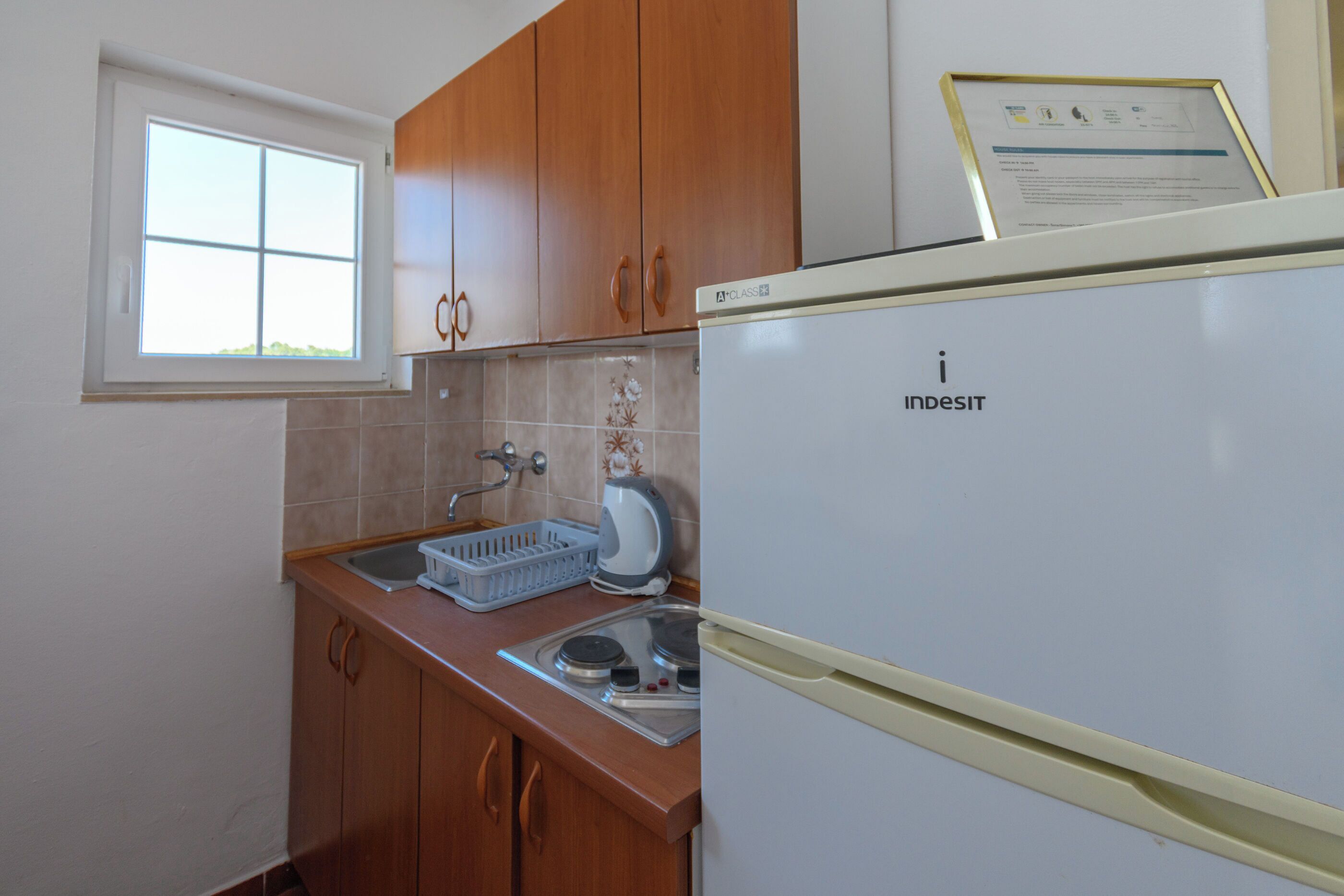 A kitchen with wooden cabinets, a window, a sink, a kettle, and a refrigerator.
