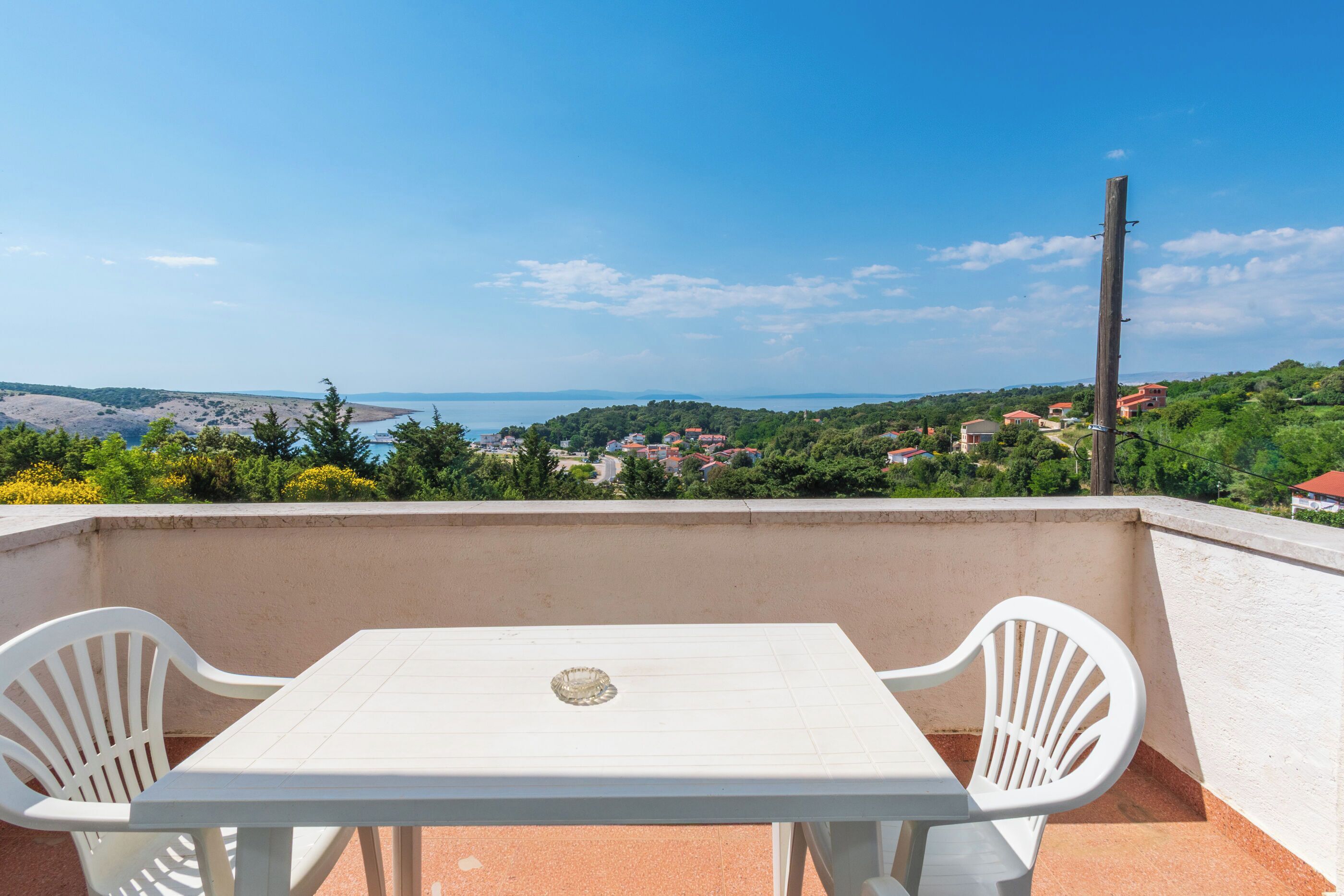 Un balcón con mesa y sillas, con vistas a un paisaje costero con casas y vegetación.