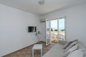Une pièce avec un canapé blanc, une petite table blanche, un téléviseur fixé au mur et une porte-fenêtre coulissante donnant sur un espace extérieur avec vue.