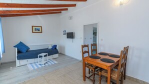 Una habitación con una cama, una mesa de comedor con sillas, una mesita auxiliar y un televisor fijado a la pared, además de una lámpara.