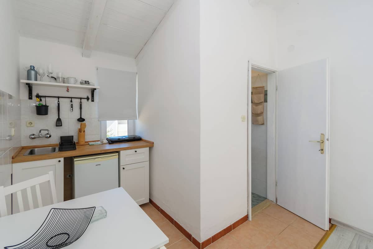Una cocina compacta con encimera blanca, un fregadero y lavavajillas. En primer plano, una mesa blanca con un mantel individual negro.