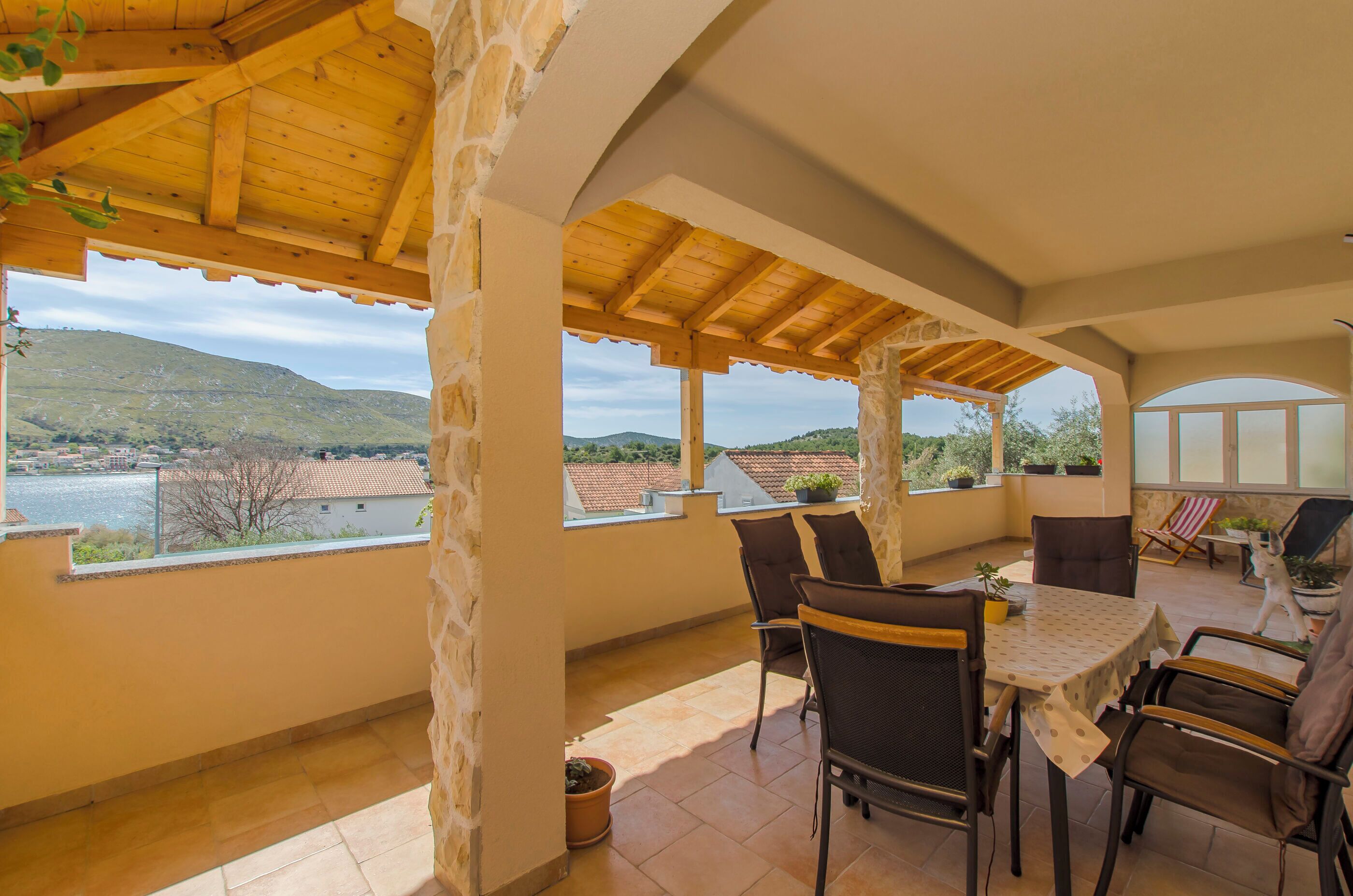 A covered patio with a table and chairs, large windows, and a view of a hillside.