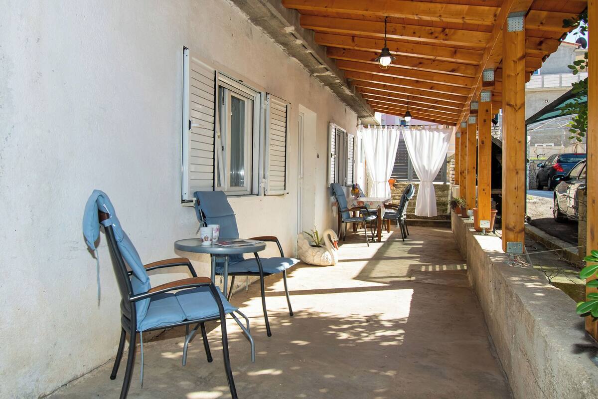An outdoor patio with wooden beams, a covered seating area with chairs and tables, and a view of parked cars in the background.