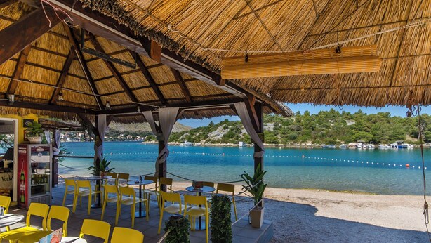 A beachside restaurant with outdoor seating, a thatched roof, and a view of the sea. - Apartments Vinka (Grebastica)