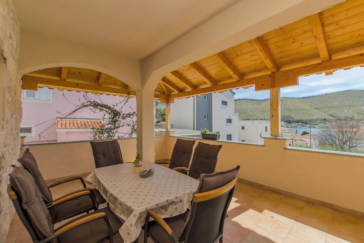 A covered outdoor dining area with a table and chairs, a potted plant, and a view of buildings and hills.