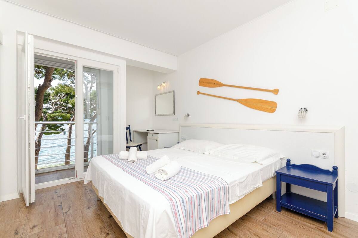 A bedroom with a bed, a blue nightstand, a desk with a chair, a window with a view of trees, and two wooden paddles on the wall.