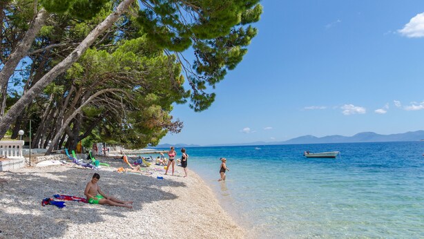 A beach with clear blue water, people sunbathing, and a boat in the distance. - Apartments Beach House Paradiso (Podgora)