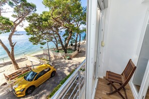 En balkong med utsikt över havet, en gul bil parkerad utanför och en trästol inomhus.
