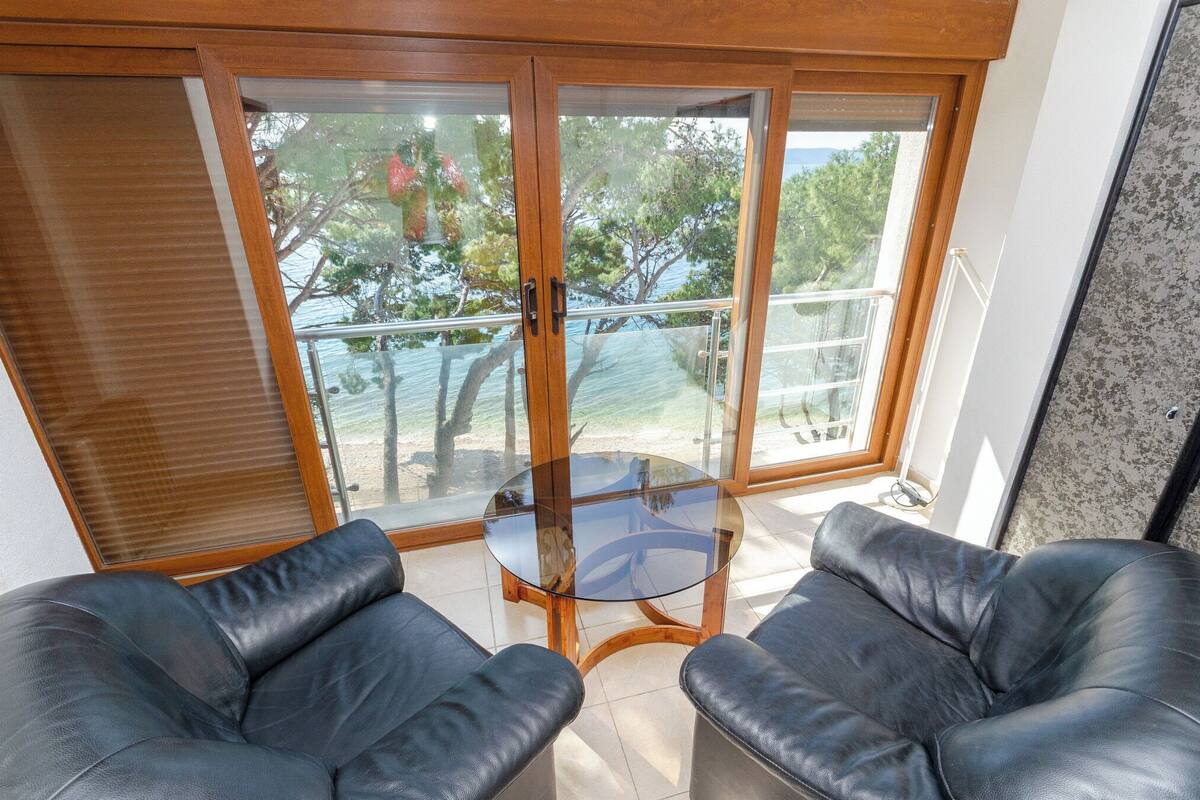 A room with a glass table, two black leather chairs, and a view through large glass doors to the outside.