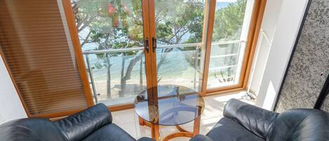 A room with a glass table, two black leather chairs, and a view through large glass doors to the outside.