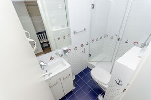 Un baño con lavamanos, inodoro y zona de ducha blancos, con azulejos de temática náutica y un espejo.