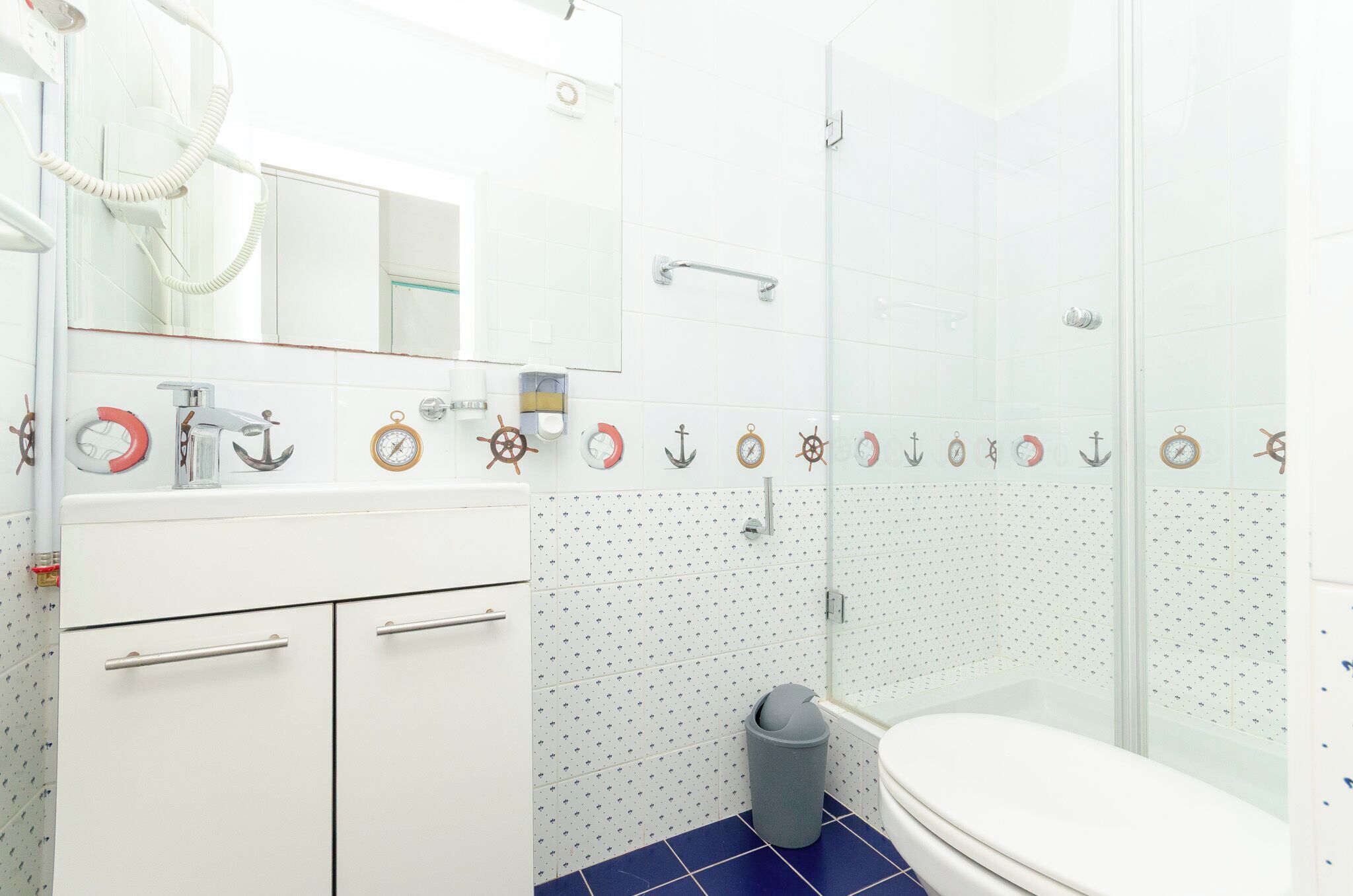 Et badeværelse med et hvidt håndvask, spejl og toilet, samt en bruseniche med glasdør og flisegulv.