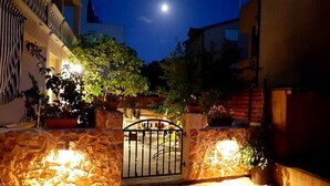 Une entrée fermée par un portail, avec des murs en pierre et des plantes en pot, qui mène à un bâtiment où l’on peut apercevoir la lune dans le ciel.