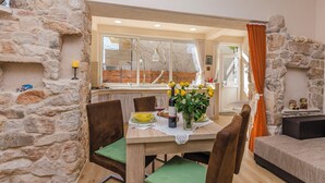 Un coin repas agrémenté d’un mur en pierre, d’une table en bois dressée pour deux personnes et d’une vue sur la cuisine grâce à une porte vitrée.