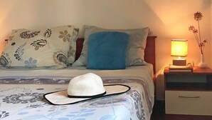 En pænt redt seng med et mønstret sengetæppe, en hvid hat på sengen og et natbord med en lampe og en vase med blomster.