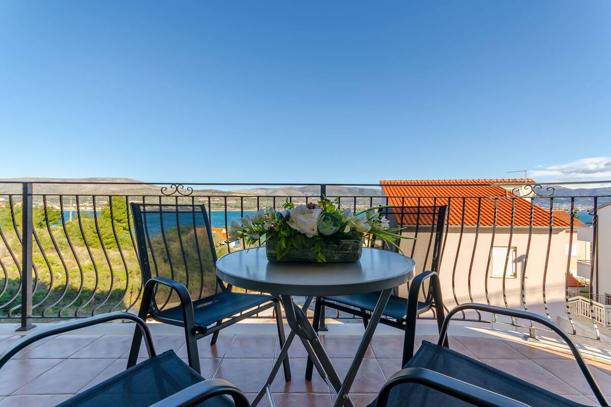 A balcony with a round table and chairs, a flower arrangement, and a view of the sea and mountains.