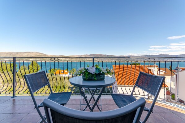 A balcony with two chairs and a table, overlooking a coastal view with mountains and a clear sky.