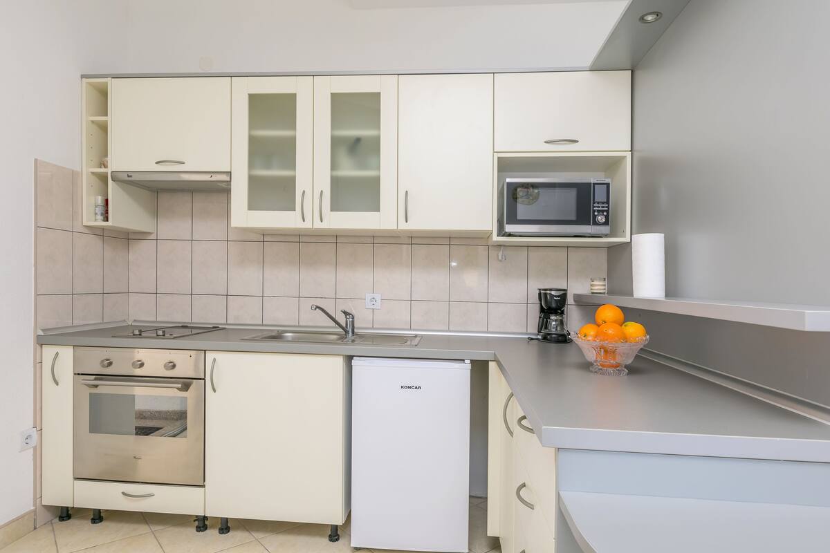 A compact kitchen with white cabinets, a built-in microwave, a small refrigerator, and a bowl of oranges on the counter.
