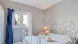 Quarto com uma cama branca, uma mesa de cabeceira com luminária e uma janela com cortinas.