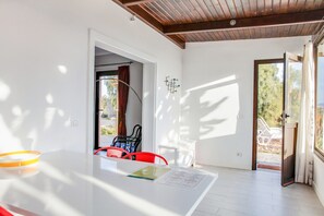 Ein heller Raum mit einem weißen Tisch, roten Stühlen, einer Holzbalkendecke und einer Tür, die nach draußen führt.