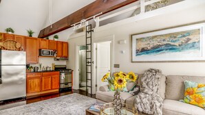 Eine Küche mit Holzausstattung, einem Kühlschrank, einer Mikrowelle und einem Herd. Eine Schiebetür führt ins Badezimmer. Im Wohnbereich befindet sich eine Couch mit einem Blumenkissen und ein Meeresblick-Gemälde.