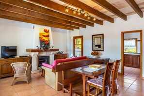 Una sala de estar con chimenea, vigas de madera en el techo, televisor, muebles de mimbre y un comedor con sillas y una mesa.