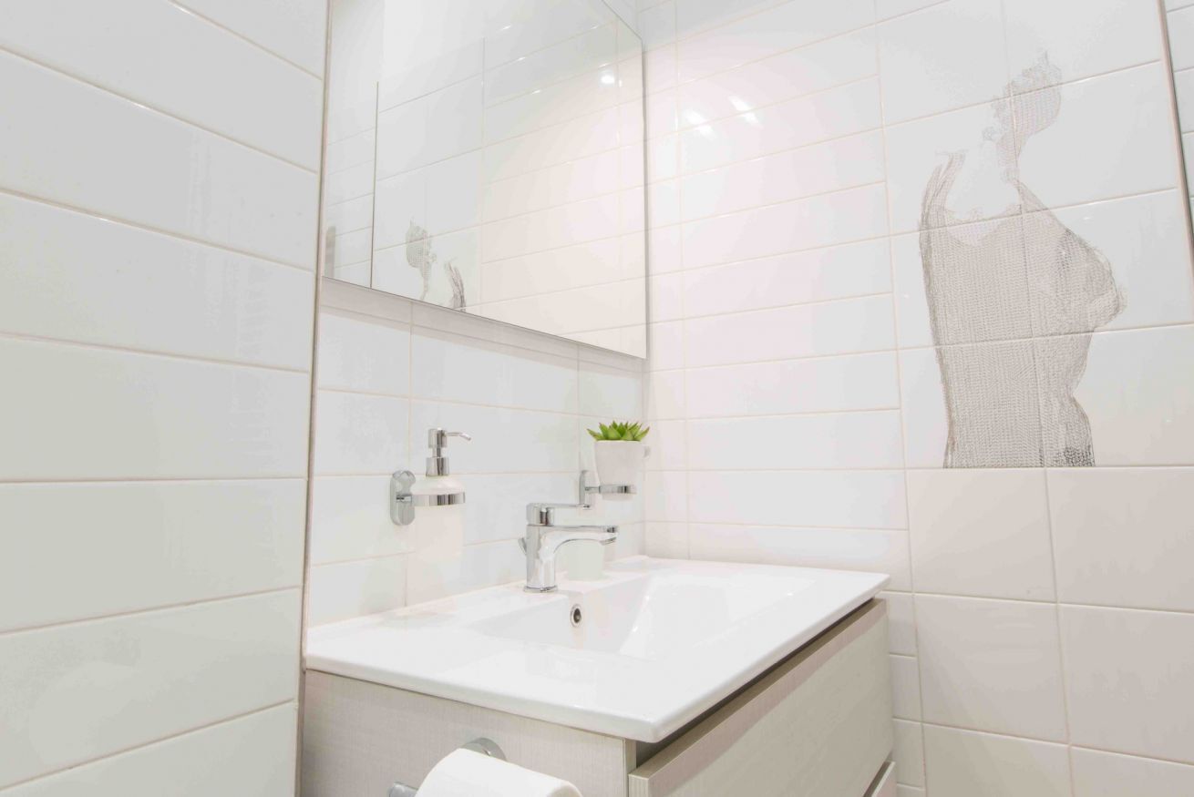 Une salle de bain moderne avec des carreaux blancs, un grand miroir, un lavabo agrémenté d’une plante et des toilettes.