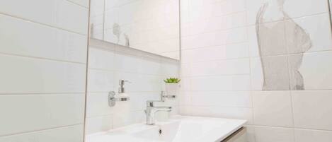 Une salle de bain moderne avec des carreaux blancs, un grand miroir, un lavabo agrémenté d’une plante et des toilettes.